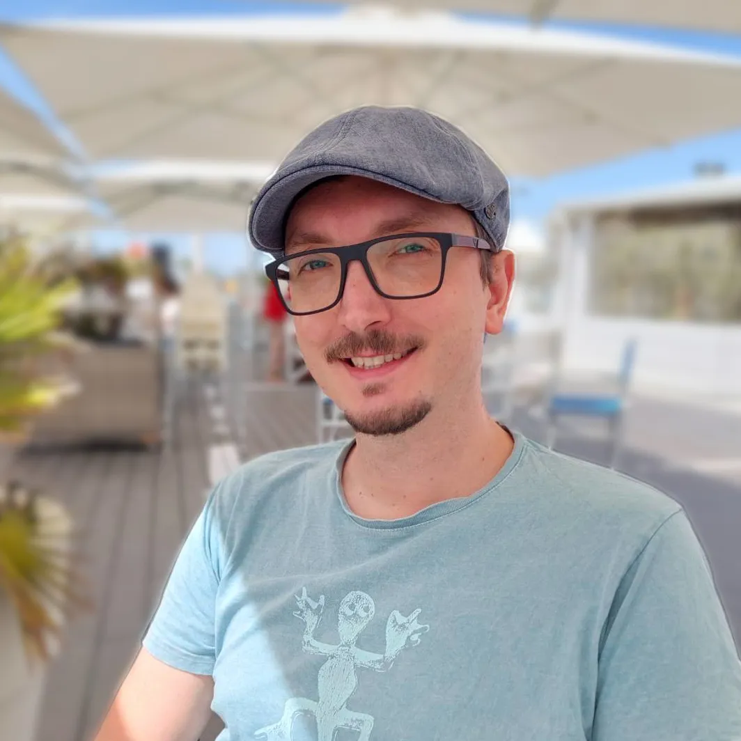 Alexander wearing a grey flat cap, dark rimmed glasses and a light blue T-shirt.