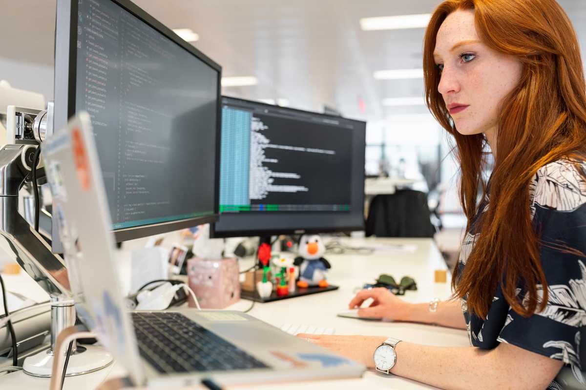 A web developer sitting at her desk, facing three monitors. Lines of code are visible on a monitor.