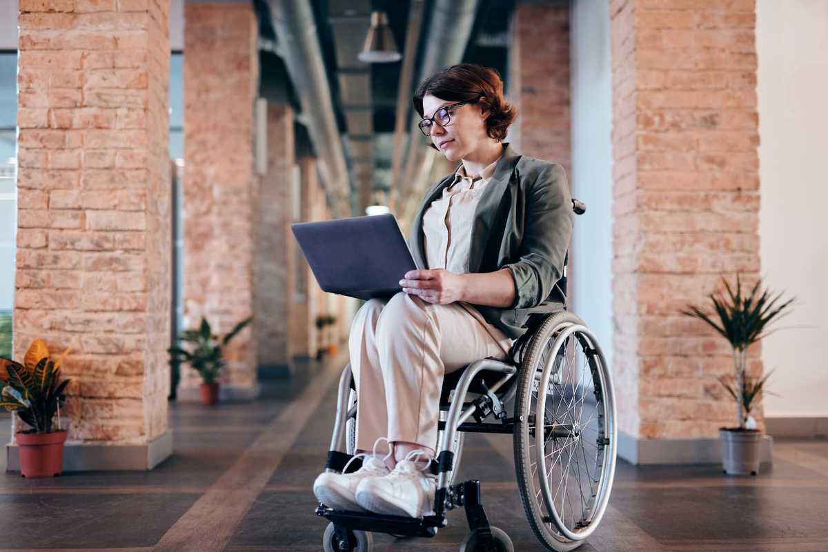 Eine Frau im Rollstuhl. Sie balanciert einen Laptop auf ihren Knien.